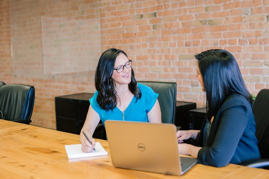 Two women discussing AI orchestration challenges at a table with a laptop open between them